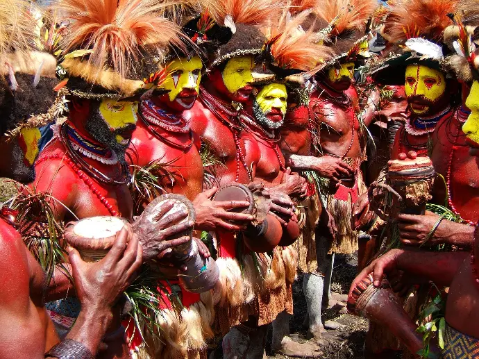 Hommes papous en parure de plumes et peinture faciale rouge et jaune jouant du tambour kundu