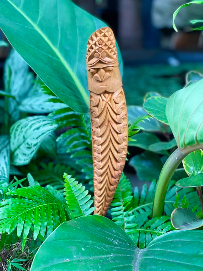 Sculpture kanak en bois clair à visage sculpté et motifs de plumes, parmi les fougères