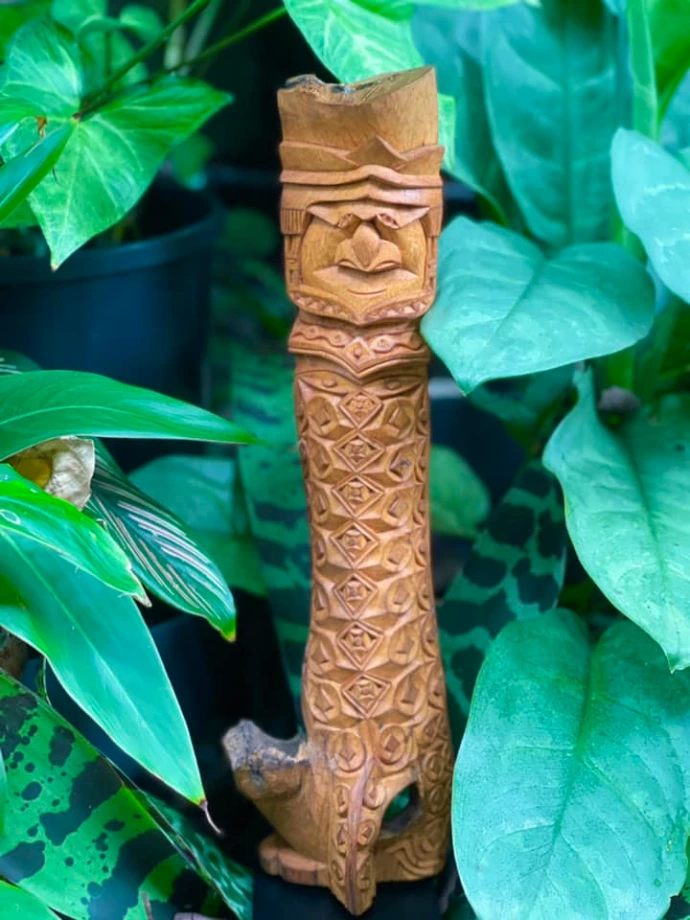 Sculpture kanak en bois à motif losangique et visage sculpté, posée dans un pot parmi les philodendrons