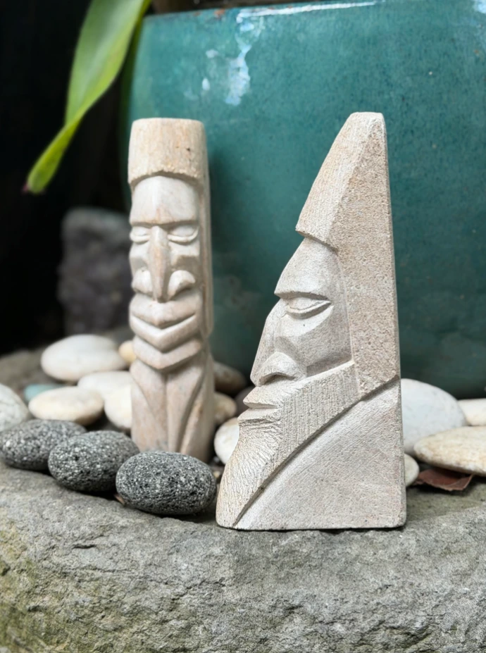 Deux sculptures en pierre à savon de Pouébo : visage anguleux barbu et fine statuette à coiffe, parmi les galets du jardin