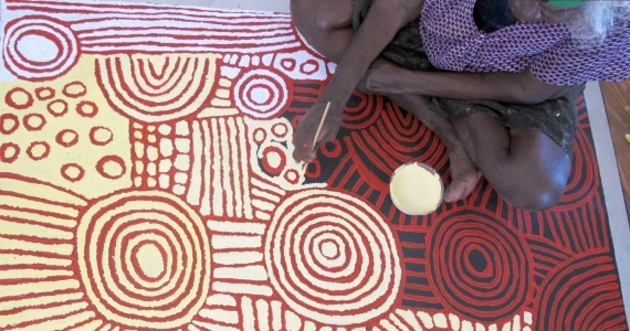 L'artiste aborigène Nungurrayi peignant au bâtonnet une toile à motifs de cercles concentriques rouge, jaune et crème