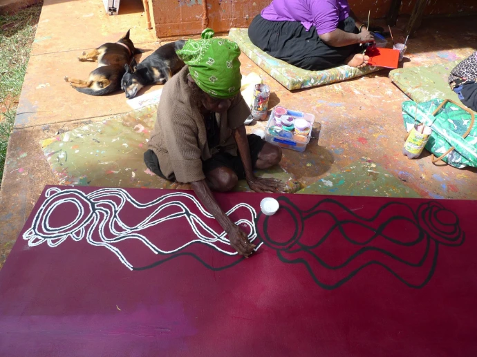 L'artiste aborigène Judy Watson peignant en extérieur, à Yuendumu, une toile bordeaux à motifs linéaires blancs et noirs