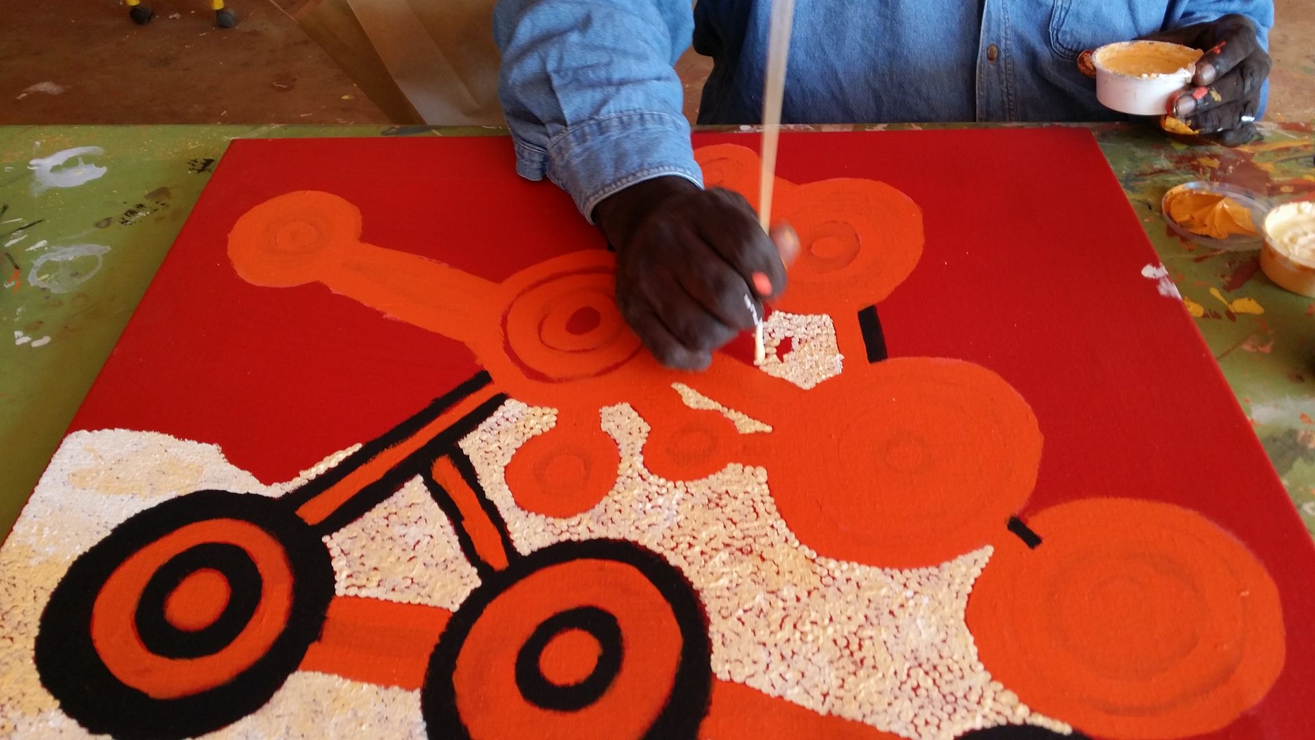 Détail de la réalisation d'une toile de l'artiste Johnny Yungut Tjupurrurla, motifs de points et cercles concentriques orange et blancs sur fond rouge