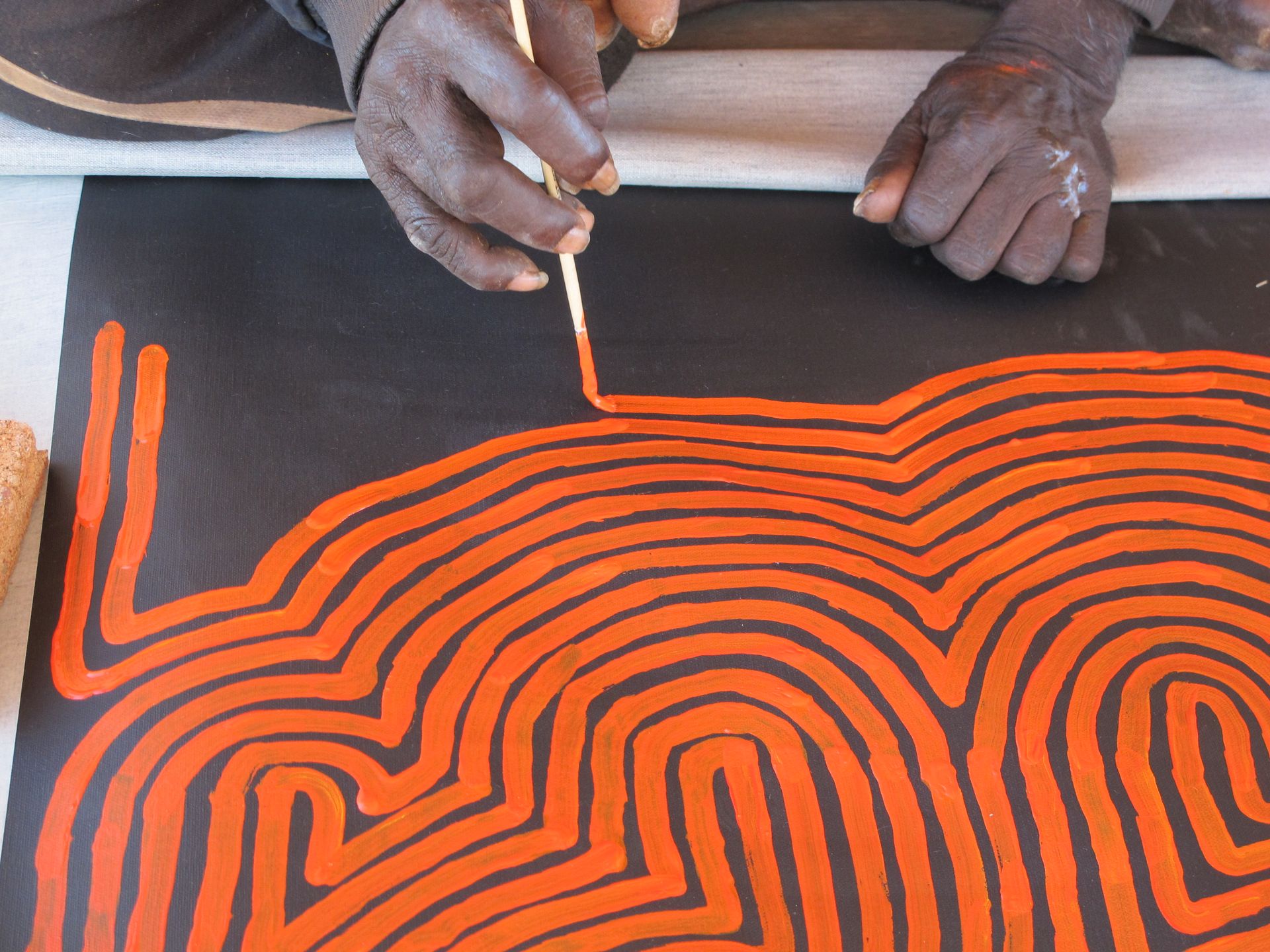 Détail de la réalisation d'une toile de l'artiste Ronnie Tjampitjinpa, motifs de lignes labyrinthiques orange sur fond noir