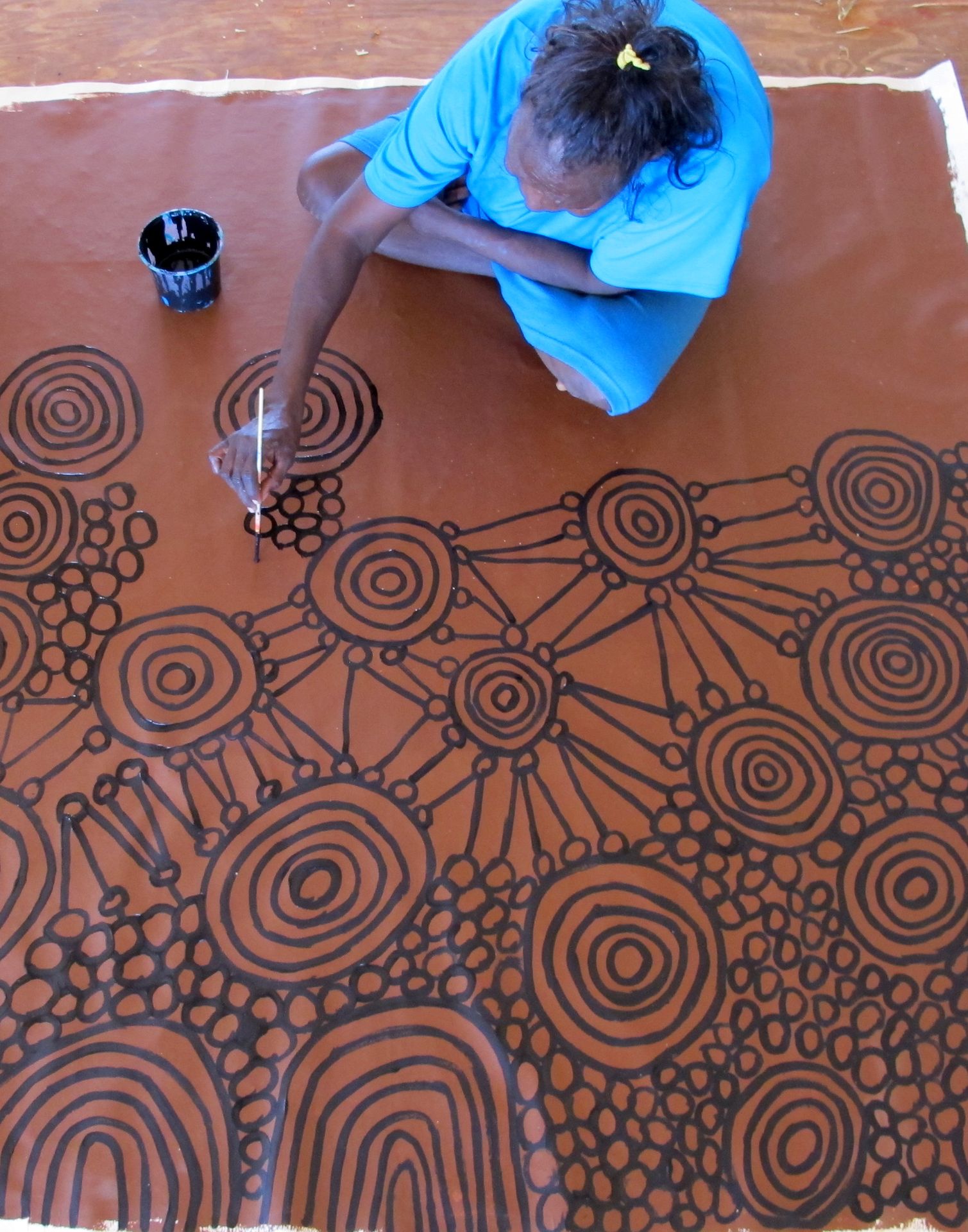 Jeune artiste aborigène australienne peignant vue de dessus une toile ocre à motifs de cercles concentriques reliés