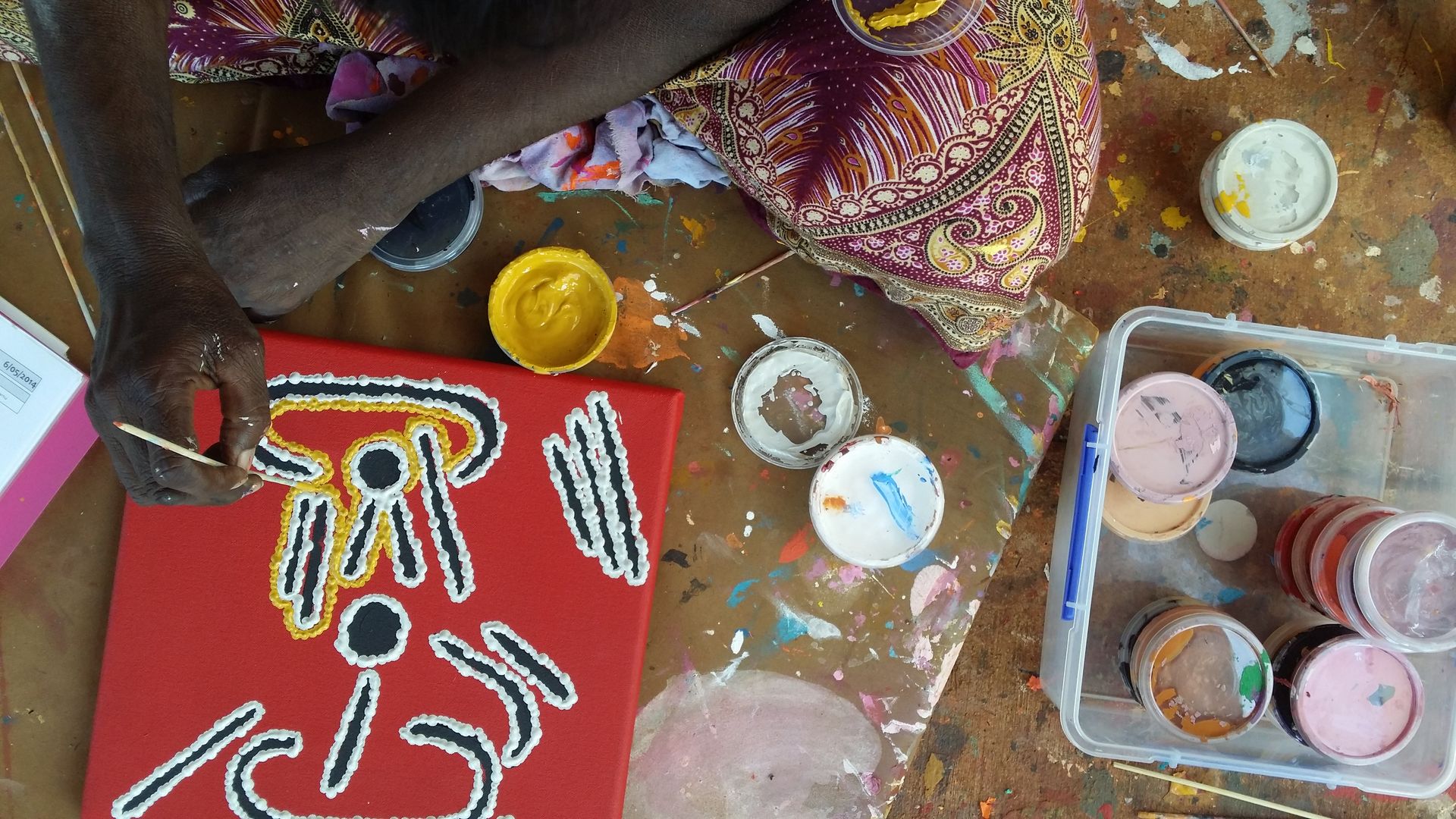 Main d'artiste aborigène peignant au bâtonnet une figure stylisée noire, blanche et jaune sur fond rouge