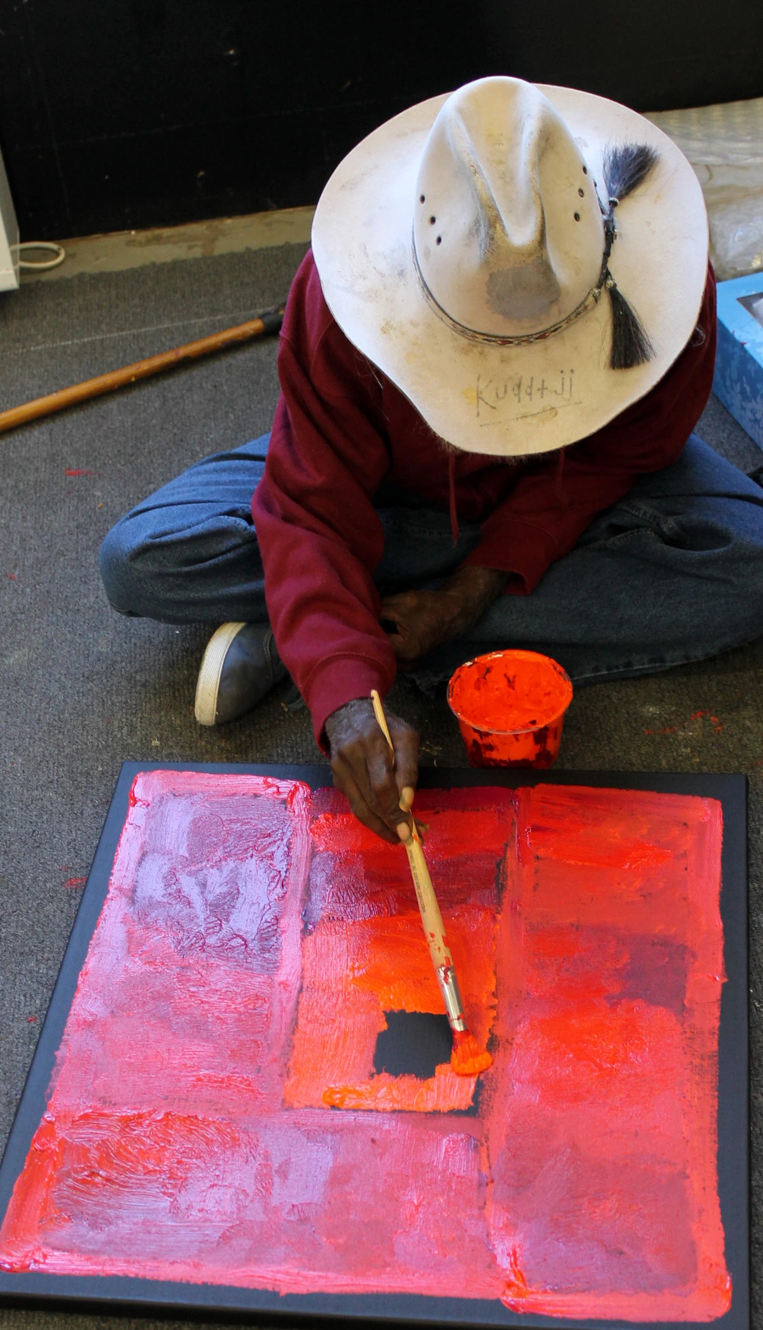 L'artiste aborigène Kudditji Kngwarreye peignant assis une toile abstraite en aplats de rouge et rose