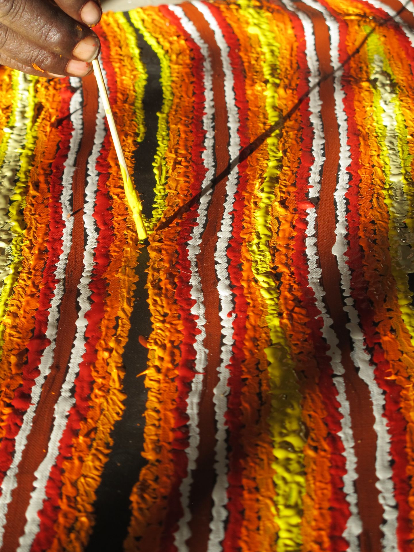 Main d'artiste aborigène peignant une œuvre d'Eileen Napaltjarri, lignes verticales striées rouge, orange, jaune et blanc
