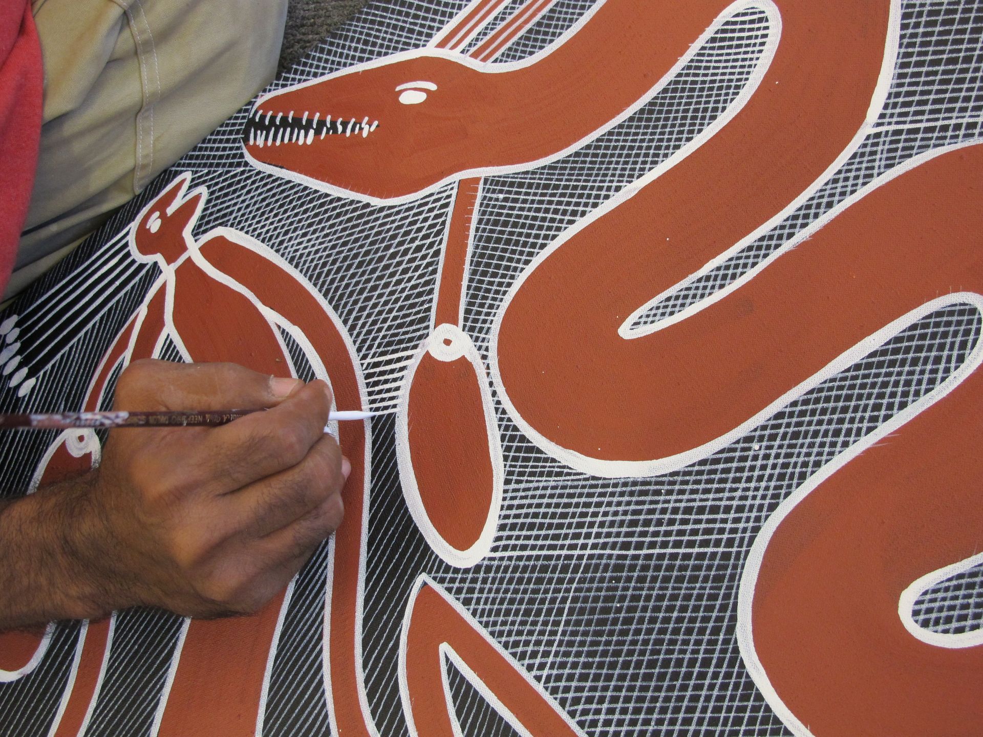 Main d'artiste aborigène peignant une œuvre d'Eddy Blitner, figure animale stylisée en hachures croisées rouille et blanc
