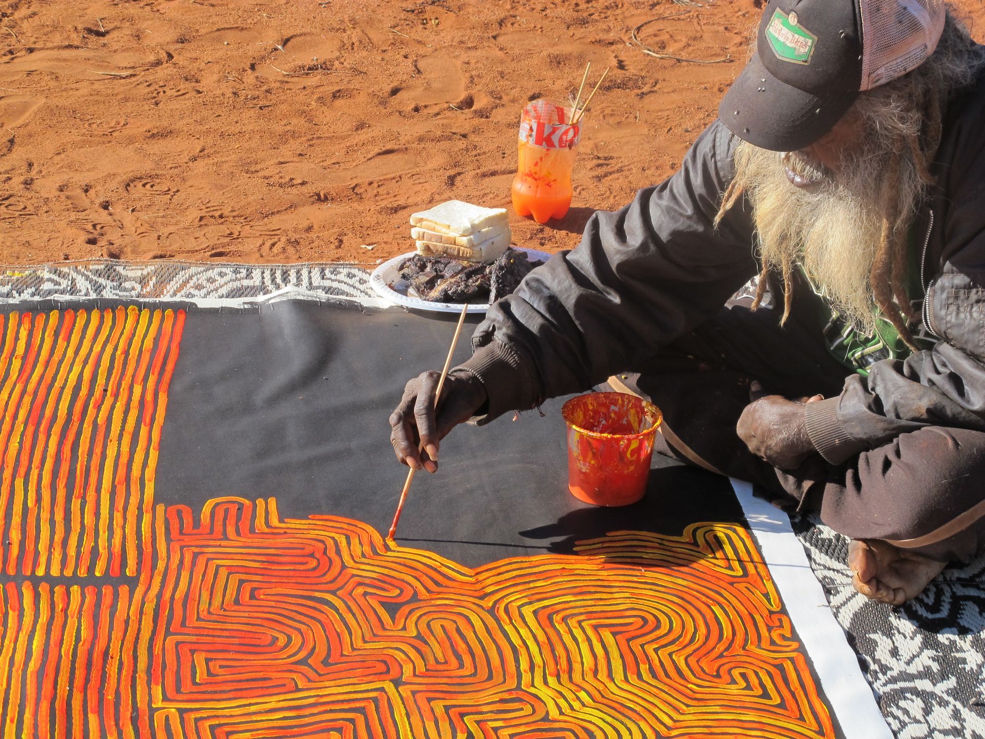 L'artiste aborigène Ronnie Tjampitjinpa peignant en plein air des lignes sinueuses orange et rouge sur fond noir