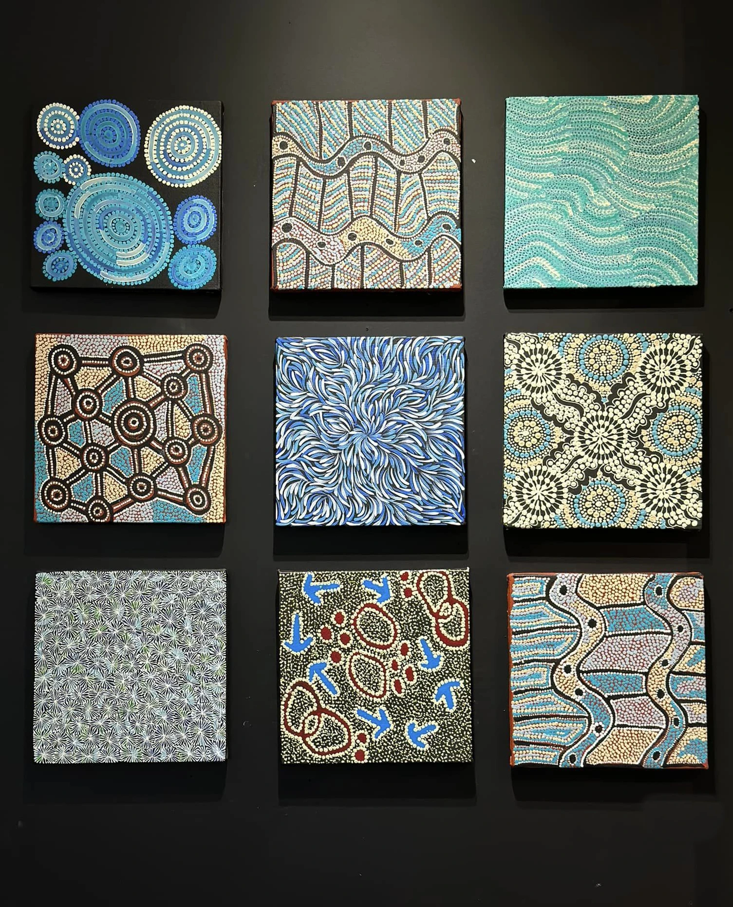 Neuf petites toiles d'art aborigène présentées en mosaïque sur un mur noir, motifs de points bleus, beige et blancs