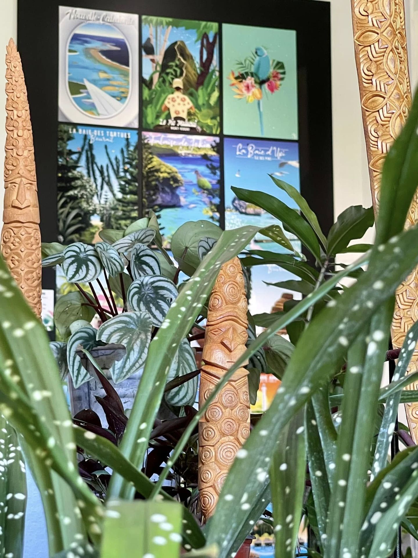 Totems kanak d'Hervé Brukoa aux visages sculptés et motifs en spirale, devant une affiche de Nouvelle-Calédonie