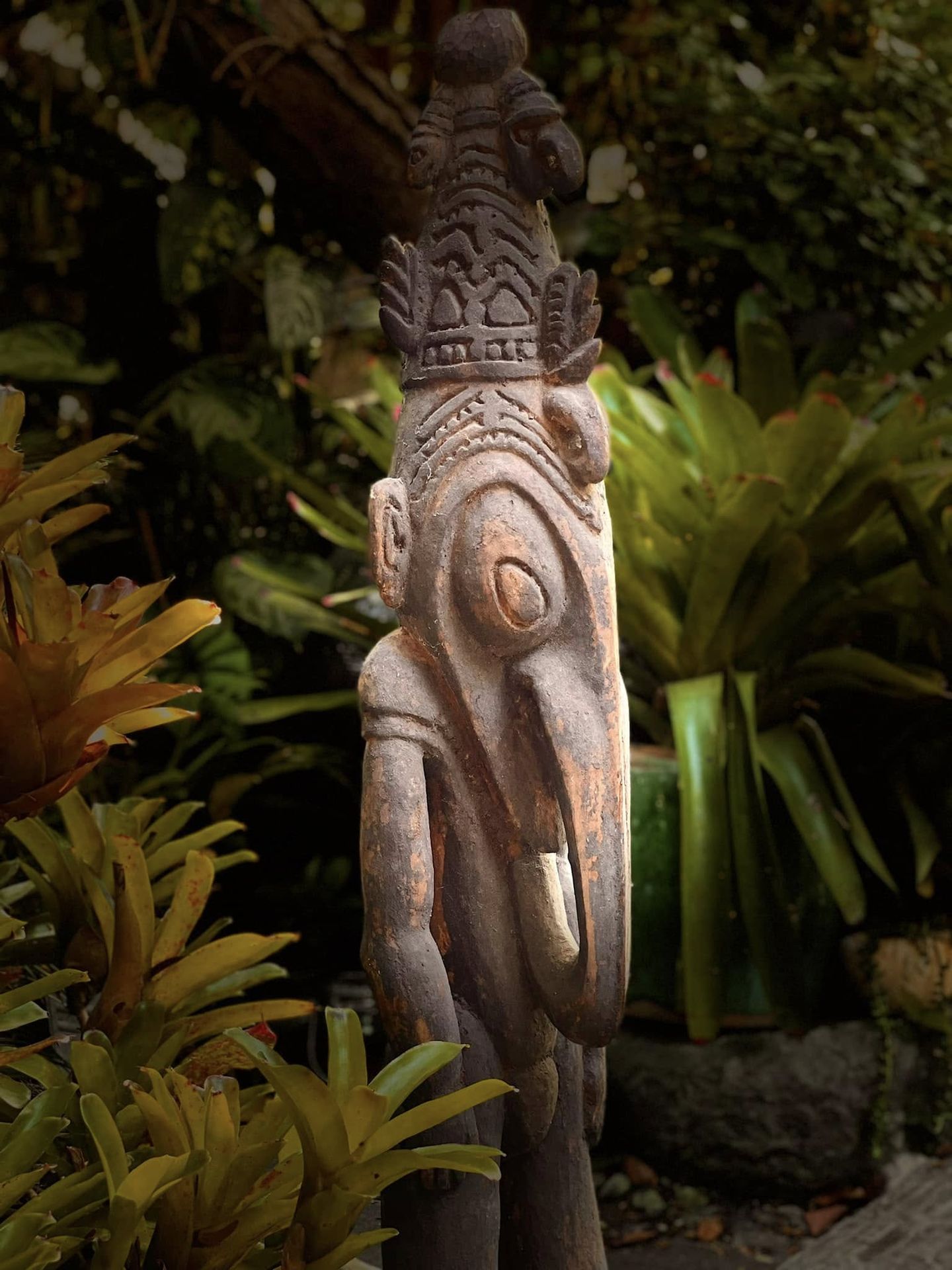 Statuette ramu représentant un homme-oiseau au bec sculpté et à la coiffe ouvragée, exposée dans un jardin tropical, exposition Drôles d'oiseaux